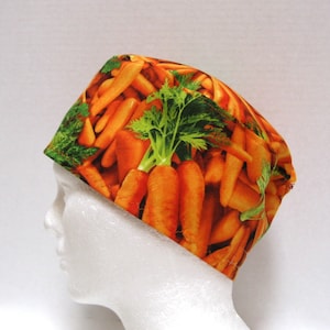 May include: A scrub cap with a bright pattern of orange carrots and green tops. The cap is made of fabric and is designed for use in medical or culinary environments. The cap is displayed on a white mannequin head.