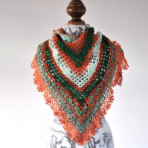 May include: A triangular crochet shawl in shades of orange, green, white, and brown. The shawl has an openwork design and is finished with a scalloped orange trim. The handmade shawl is displayed on a mannequin. The measurements are approximately 150 cm across.