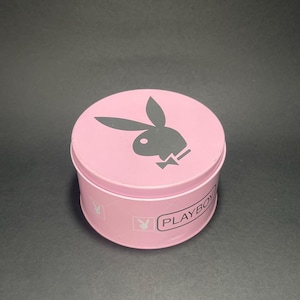 Könnte beinhalten: Eine runde, hellrosa Metalldose mit einem dunkelgrauen Playboy-Hasenlogo auf dem Deckel. Die Seite des Behälters zeigt das Wort PLAYBOY in einem rechteckigen Rahmen, begleitet von einem sich wiederholenden Muster aus kleinen Hasensymbolen und Linien.