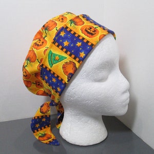 May include: A Halloween-themed scrub cap featuring a repeating pattern of orange pumpkins on a yellow background. The cap has a blue border with gold stars and a green witch hat design. The cap is tied at the back.