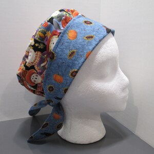 May include: A reversible head covering with a blue and fall-themed pattern. One side features pumpkins and sunflowers on a blue background, while the other has scarecrows and autumn leaves. The head covering is tied at the back.