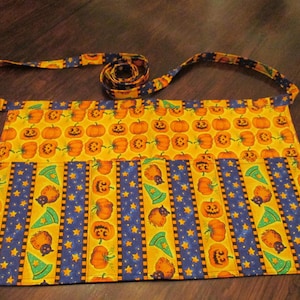 May include: A rectangular waist apron featuring a vibrant Halloween pattern with orange jack-o'-lanterns, green witch hats, and black cats on a yellow background, interspersed with vertical blue stripes decorated with yellow stars and a matching strap.
