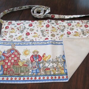 May include: A white and cream kitchen apron with a colorful farm scene print. The apron has a long tie and a large pocket on the front. The print features a red barn, a farmer, chickens, and apples.
