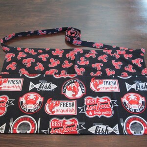 May include: Black apron with a red lobster and seafood design. The apron features various labels with text such as "Fresh Crawfish", "Best Seafood", and "Fish". The apron has a black strap with a red lobster pattern.