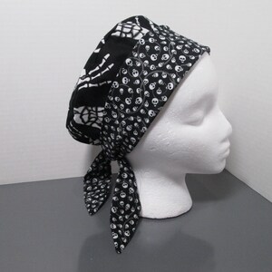 May include: A black and white scrub cap featuring a pattern of skeletal hands on the main section and a band decorated with small skulls. The cap includes two fabric ties at the back, constructed from patterned cotton fabric with a fitted design.