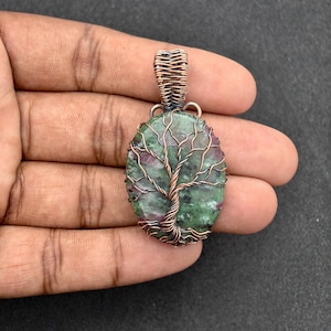 Può includere: Ciondolo ovale con una pietra verde e rossa, con un design ad albero in filo di rame. Il ciondolo ha un'ansa in filo di rame ed è lungo circa 3,8 cm.