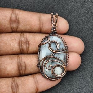 Peut inclure: Pendentif ovale avec une pierre blanche et grise, envelopp&eacute;e de fil de cuivre. Le fil est con&ccedil;u avec des volutes et des boucles, cr&eacute;ant un aspect unique et artistique. Le pendentif est suspendu &agrave; une cha&icirc;ne en cuivre.
