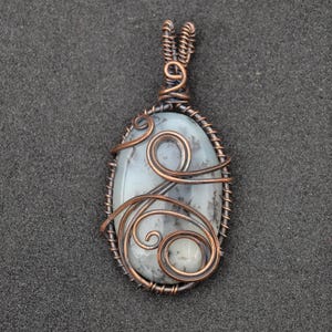 Peut inclure: Pendentif ovale avec une pierre gris clair vein&eacute;e de gris fonc&eacute;, envelopp&eacute;e de fil de cuivre. Le fil de fer comprend des volutes d&eacute;coratives et une b&eacute;li&egrave;re pour un collier. Bijou artisanal.