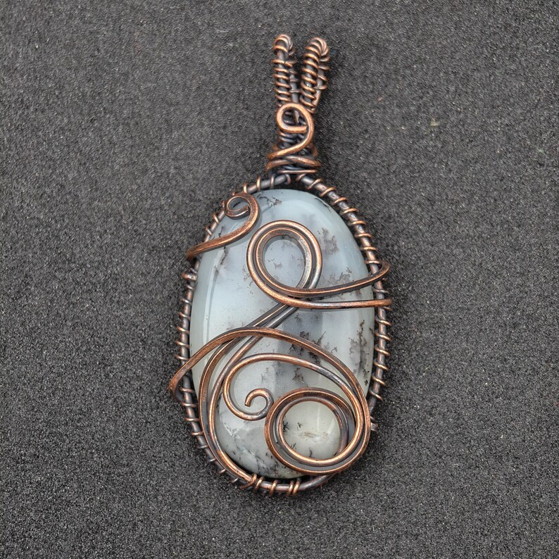 Peut inclure: Pendentif ovale avec une pierre gris clair vein&eacute;e de gris fonc&eacute;, envelopp&eacute;e de fil de cuivre. Le fil de fer comprend des volutes d&eacute;coratives et une b&eacute;li&egrave;re pour un collier. Bijou artisanal.