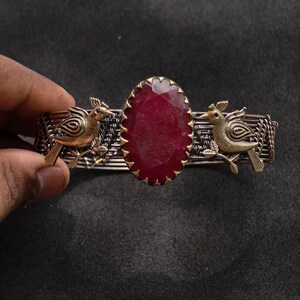 May include: A handcrafted bracelet featuring a large, oval, deep red gemstone set in a gold-toned bezel. Two gold-colored bird figures flank the gemstone, and the bracelet band has a woven metal design. The bracelet is held by a hand.