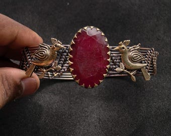 May include: A handcrafted bracelet with a large, oval, deep red gemstone set in a gold-toned bezel. Two gold-coloured bird figures flank the gemstone, and the bracelet band has a woven metal design. The bracelet is held by a hand.