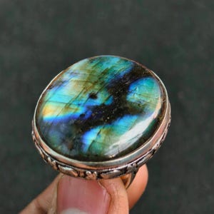 Può includere: Anello ovale con pietra di labradorite. La pietra mostra sfumature iridescenti blu, verdi e dorate. L'anello ha una fascia color argento con un design decorativo attorno alla pietra.
