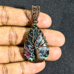 May include: A teardrop-shaped pendant with a dark green and red stone, wrapped in copper wire. The wire forms a tree design, enhancing the natural aesthetic of the jewellery. The pendant is held in a hand.