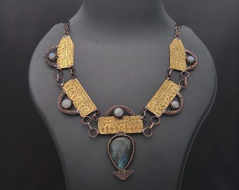 May include: A statement necklace with a distinctive design. It features gold-coloured rectangular and trapezoid-shaped metal pieces with detailed patterns, linked by copper-coloured chains. A teardrop-shaped pendant with a blue-green stone and small, round, white stones completes the piece.