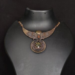 May include: An ornate copper necklace with a detailed design. The necklace features a central pendant with a large, green gemstone and a circular wireframe. The upper section has a wing-like shape with embossed patterns and a small, orange stone.