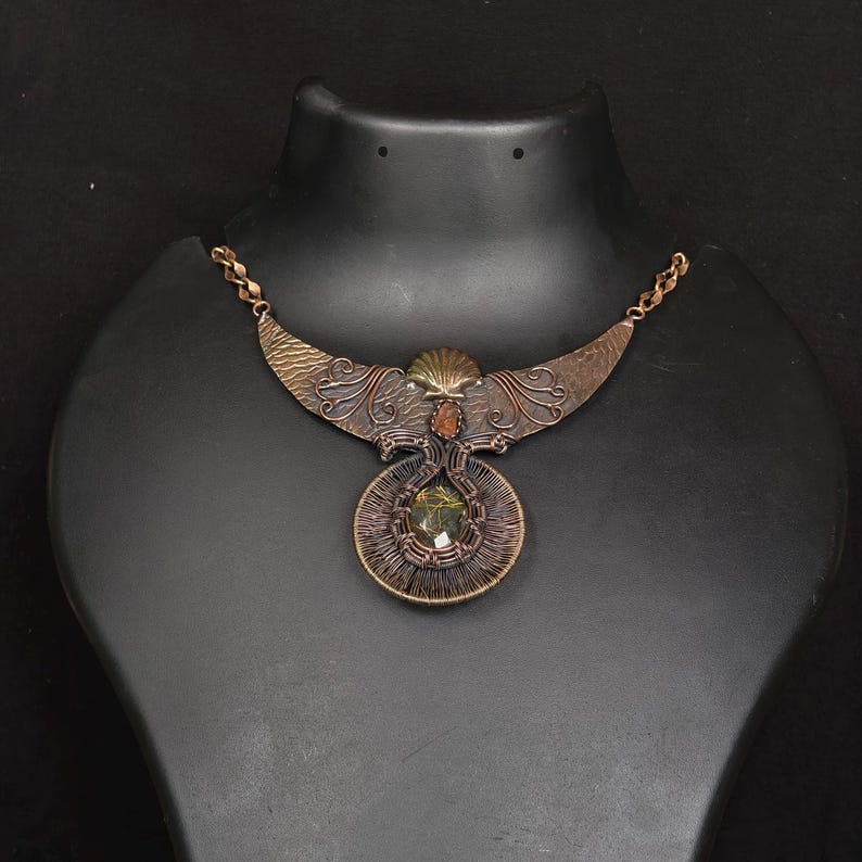 May include: An ornate copper necklace with a detailed design. The necklace features a central pendant with a large, green gemstone and a circular wireframe. The upper section has a wing-like shape with embossed patterns and a small, orange stone.
