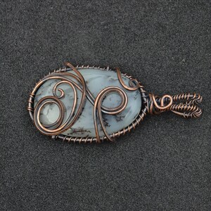 Peut inclure: Pendentif ovale avec une pierre bleu-gris p&acirc;le, orn&eacute;e d'inclusions sombres, envelopp&eacute;e de fil de cuivre. Le filigrane comprend des volutes d&eacute;coratives et une b&eacute;li&egrave;re boucl&eacute;e. Le pendentif est pos&eacute; sur un fond gris fonc&eacute;.