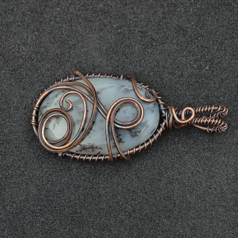 Peut inclure: Pendentif ovale avec une pierre bleu-gris p&acirc;le, orn&eacute;e d'inclusions sombres, envelopp&eacute;e de fil de cuivre. Le filigrane comprend des volutes d&eacute;coratives et une b&eacute;li&egrave;re boucl&eacute;e. Le pendentif est pos&eacute; sur un fond gris fonc&eacute;.