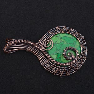May include: A handcrafted pendant featuring a vibrant green stone, intricately wrapped in copper wire. The wirework forms a decorative frame and bail, with spiral accents. The pendant is displayed against a black background, highlighting the contrast of colors and textures.