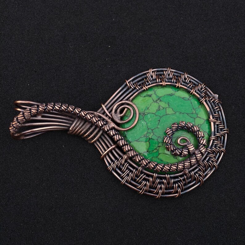 May include: A handcrafted pendant featuring a vibrant green stone, intricately wrapped in copper wire. The wirework forms a decorative frame and bail, with spiral accents. The pendant is displayed against a black background, highlighting the contrast of colors and textures.
