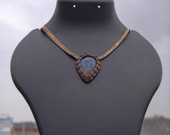 May include: A teardrop-shaped labradorite pendant featuring a blue iridescent flash, encased in intricate copper wire wrapping. The pendant is attached to a flat, woven copper chain that extends symmetrically from the top of the setting.