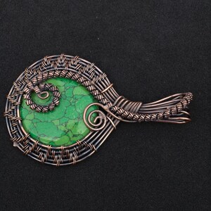 May include: A handcrafted pendant featuring a vibrant green stone, intricately wrapped with copper-colored wire. The wirework forms a decorative pattern around the stone, with a spiral design at the top. The pendant has a unique, organic shape.