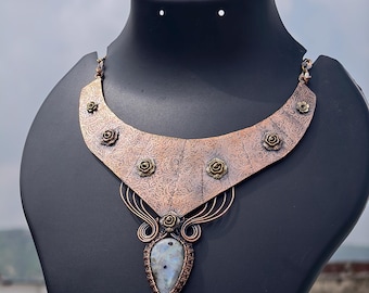 May include: A copper-toned statement necklace with a floral design and a teardrop-shaped stone pendant. The necklace features intricate metalwork and is displayed on a black mannequin. The necklace is a unique piece of jewelry.