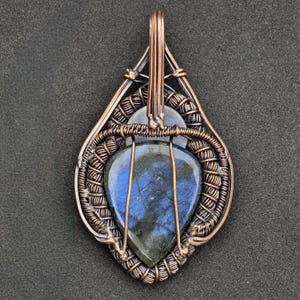 Peut inclure: Pendentif artisanal en fil de cuivre avec une pierre de labradorite en forme de goutte d'eau, aux reflets bleus et verts. La pierre est sertie dans un travail de filigrane complexe, cr&eacute;ant un design artistique unique. Le pendentif a une boucle pour une cha&icirc;ne.