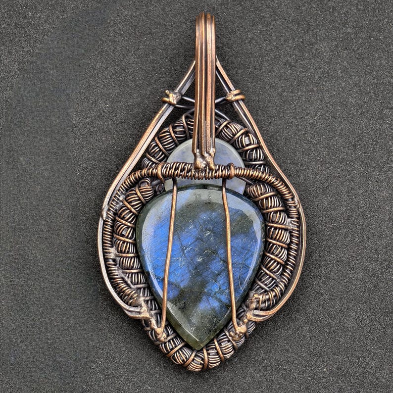 Peut inclure: Pendentif artisanal en fil de cuivre avec une pierre de labradorite en forme de goutte d'eau, aux reflets bleus et verts. La pierre est sertie dans un travail de filigrane complexe, cr&eacute;ant un design artistique unique. Le pendentif a une boucle pour une cha&icirc;ne.