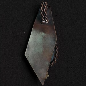 May include: A handcrafted pendant with a geometric design. The pendant features a diamond-shaped, tarnished metal plate with a braided copper wire accent. A small, decorative element is visible at the bottom. The pendant hangs from a copper wire loop.