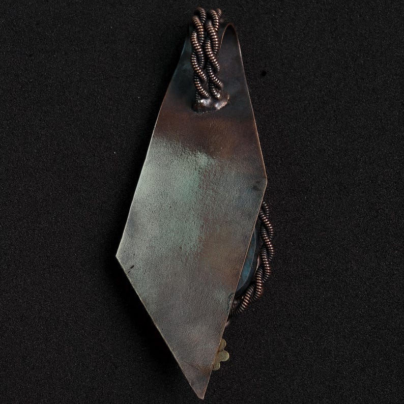 May include: A handcrafted pendant with a geometric design. The pendant features a diamond-shaped, tarnished metal plate with a braided copper wire accent. A small, decorative element is visible at the bottom. The pendant hangs from a copper wire loop.