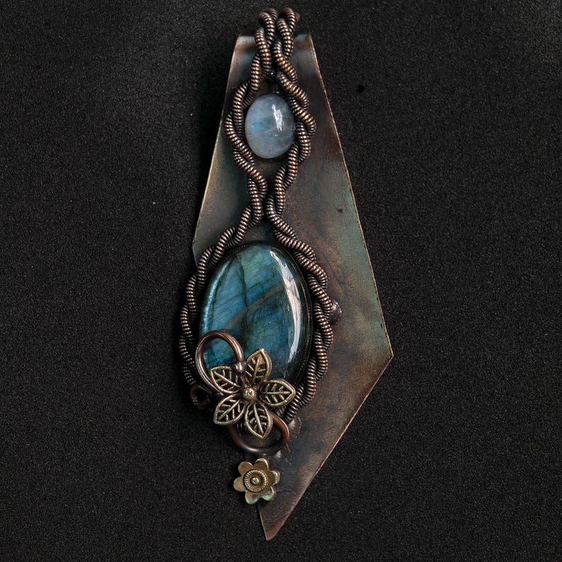May include: Handmade pendant with a large, oval labradorite stone, displaying blue and green iridescence, and a smaller, round stone. The stones are set in a copper wire wrap with a floral detail, on a diamond-shaped metal backing.