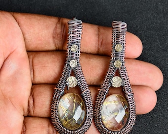 May include: A pair of handcrafted wire-wrapped earrings. The earrings feature a teardrop shape with a clear, faceted gemstone at the center. The wire is a copper color, and the earrings have small, decorative metal accents.