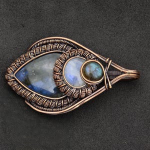 Peut inclure: Pendentif artisanal en fil de cuivre avec trois pierres pr&eacute;cieuses. Le pendentif en forme de goutte d'eau pr&eacute;sente une grande pierre bleu-gris fonc&eacute;, une petite pierre ronde bleu clair et une petite pierre ronde bleu-vert fonc&eacute;. Le filigrane est complexe.