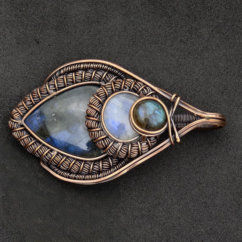 Peut inclure: Pendentif artisanal en fil de cuivre avec trois pierres pr&eacute;cieuses. Le pendentif en forme de goutte d'eau pr&eacute;sente une grande pierre bleu-gris fonc&eacute;, une petite pierre ronde bleu clair et une petite pierre ronde bleu-vert fonc&eacute;. Le filigrane est complexe.