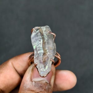 May include: A raw quartz crystal ring with copper wire detailing. The clear quartz has an iridescent sheen and is set on a copper band. The ring is displayed against a dark backdrop.