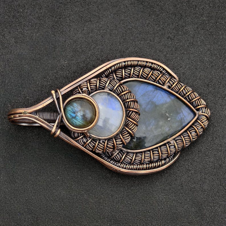 Peut inclure: Pendentif en fil de cuivre avec trois pierres pr&eacute;cieuses : une petite pierre ronde, une plus grande pierre ronde et une pierre en forme de goutte. Les pierres ont des reflets bleus et iris&eacute;s. Le pendentif est pos&eacute; sur un fond sombre.