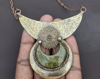May include: A necklace featuring a crescent-shaped brass pendant with an intricate floral pattern. A circular brass element with a central flower design sits above a polished green stone crescent, framed by twisted copper wire on a copper chain.