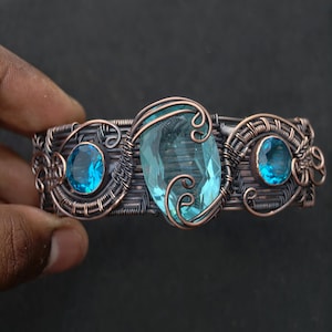 May include: A handcrafted copper wire bracelet featuring three blue gemstones. The central gemstone is an oval shape, flanked by two smaller, round stones. The wirework has intricate swirls and patterns.