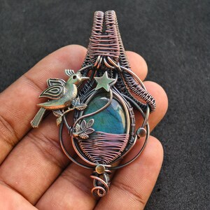 Puede incluir: Colgante artesanal con un dise&ntilde;o de alambre enrollado. El colgante presenta una piedra de color turquesa, un p&aacute;jaro de color bronce y una estrella verde. El alambre es de color cobre. El colgante se sostiene en una mano.