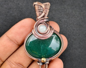 May include: A handcrafted pendant featuring a polished, oval-shaped green stone. The stone is encased in intricate copper wire, with a small, round, light-colored stone accent. The pendant has a loop for a chain.