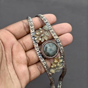 May include: A metallic bracelet featuring a braided chain design with a central circular dark gray stone setting. The piece is accented by two gold-toned floral motifs, each consisting of small leaves and berries, positioned above and below the stone.