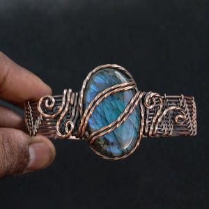 May include: Copper wire cuff bracelet featuring a large, oval labradorite stone with blue and green iridescence. The stone is secured with twisted copper wire, and the bracelet has intricate woven and coiled details.
