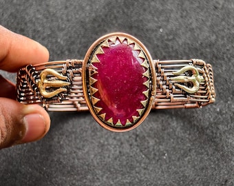 May include: A handcrafted copper wire bracelet with a large, oval, deep red gemstone at the centre. The gemstone is framed by a gold-coloured, sawtooth-edged bezel. The bracelet has gold-coloured trident accents.