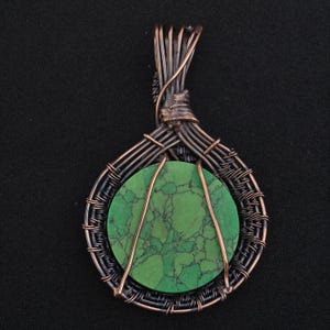 May include: Handmade pendant featuring a round, green stone with dark veining, encased in a wire-wrapped design. The pendant has a copper and black wire frame, creating a unique, artistic look. The wire is intricately woven around the stone.