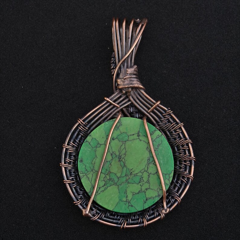 May include: Handmade pendant featuring a round, green stone with dark veining, encased in a wire-wrapped design. The pendant has a copper and black wire frame, creating a unique, artistic look. The wire is intricately woven around the stone.