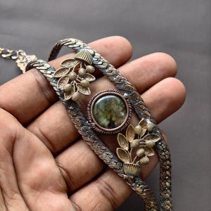 May include: A bracelet featuring a central round labradorite gemstone set in a copper-toned bezel, flanked by two metallic leaf and berry motifs. The band consists of two braided metallic strands with a dark, oxidized finish and texture.