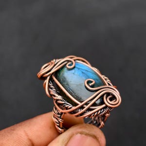 Könnte beinhalten: Handgefertigter Ring mit einem großen, ovalförmigen blau-grünen Edelstein. Der Stein ist in aufwendigem Kupferdraht gefasst, mit dekorativen Wirbeln und gewebten Mustern. Der Ring hat eine rustikale, handwerkliche Ästhetik.