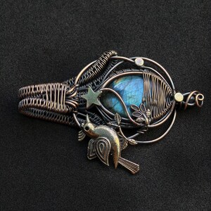 Puede incluir: Colgante artesanal con una piedra de labradorita azul, envuelta en alambre con un dise&ntilde;o detallado. El colgante incluye una estrella y un p&aacute;jaro, con tonos bronce y plata. El dise&ntilde;o general es intrincado y art&iacute;stico.