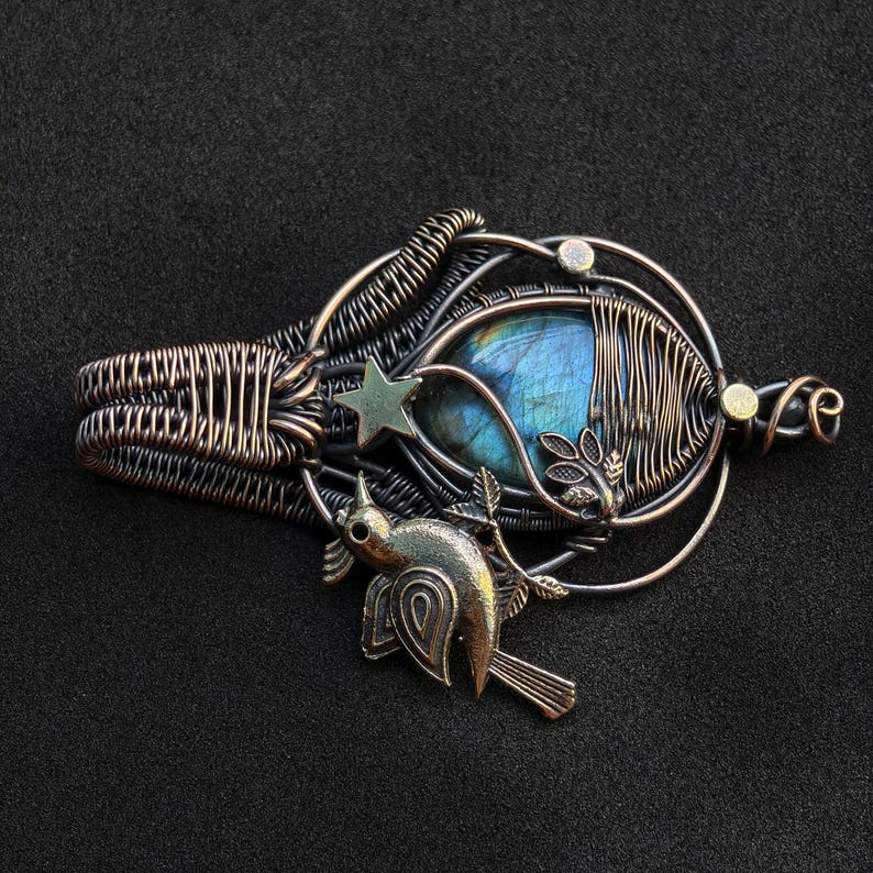 Puede incluir: Colgante artesanal con una piedra de labradorita azul, envuelta en alambre con un dise&ntilde;o detallado. El colgante incluye una estrella y un p&aacute;jaro, con tonos bronce y plata. El dise&ntilde;o general es intrincado y art&iacute;stico.